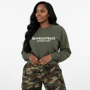 Woman wearing an olive green sweatshirt with text BEWRIGHTBACK BOARDING SOON and camouflage pants on a white background