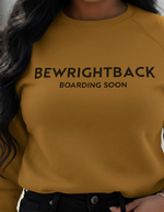 Person wearing a mustard yellow sweatshirt with text 'BEWRIGHTBACK BOARDING SOON' on a blurred background